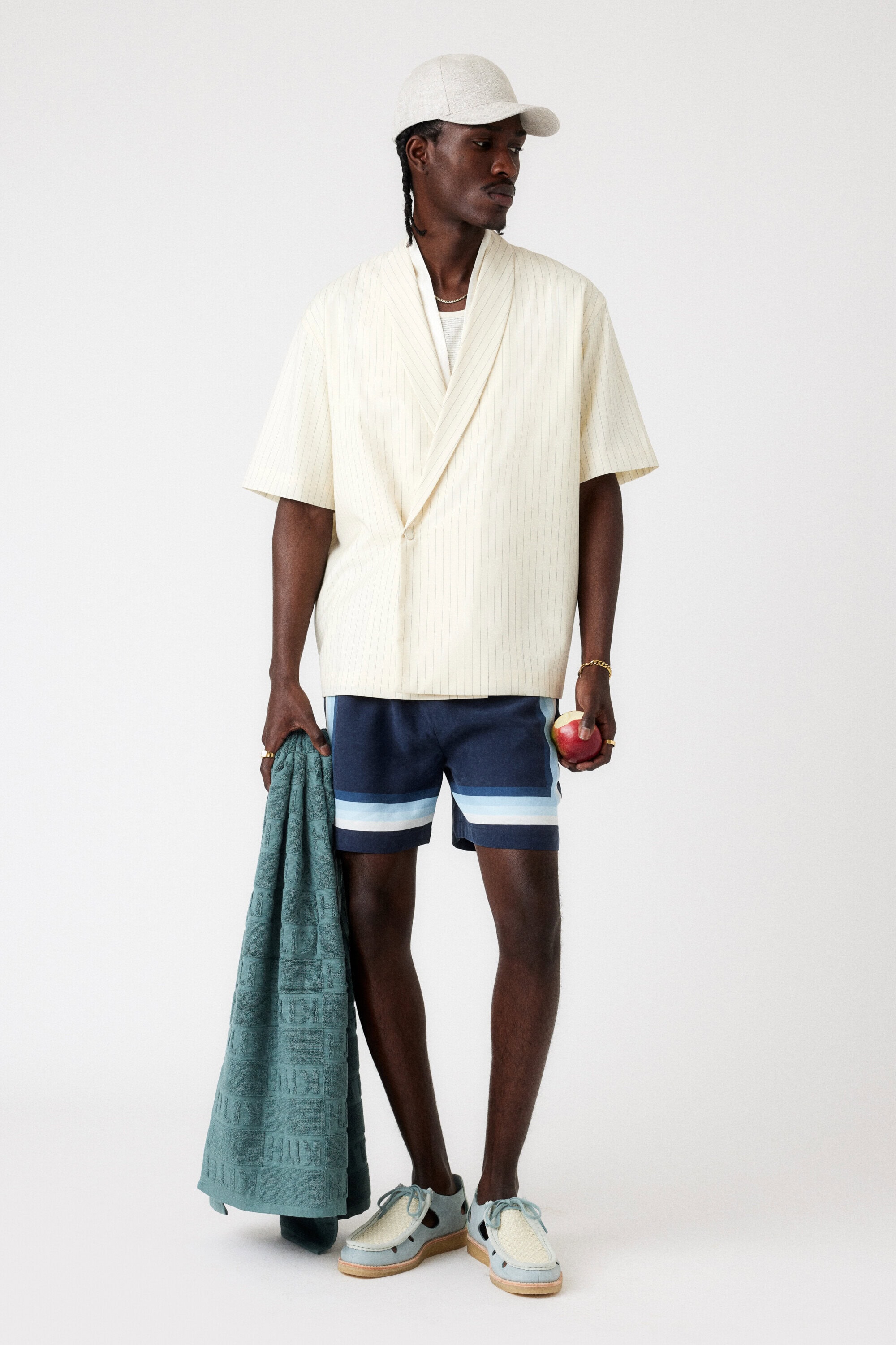 Male fashion model in a beige kimono shirt, blue striped shorts, beige cap, and matching shoes, standing in a white studio.