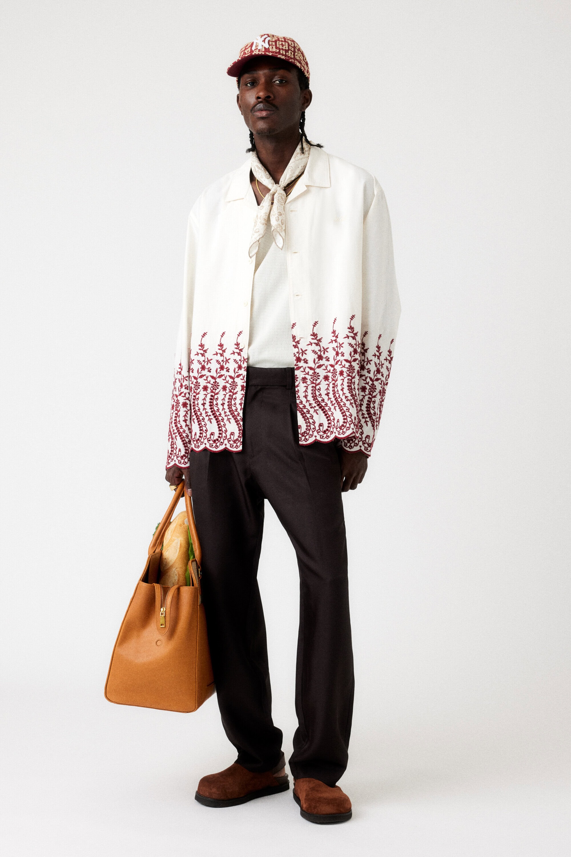 Full-length studio shot of a Black male model wearing a white button-down shirt with brown trim, dark pants, cap, and carrying a tan leather tote bag.