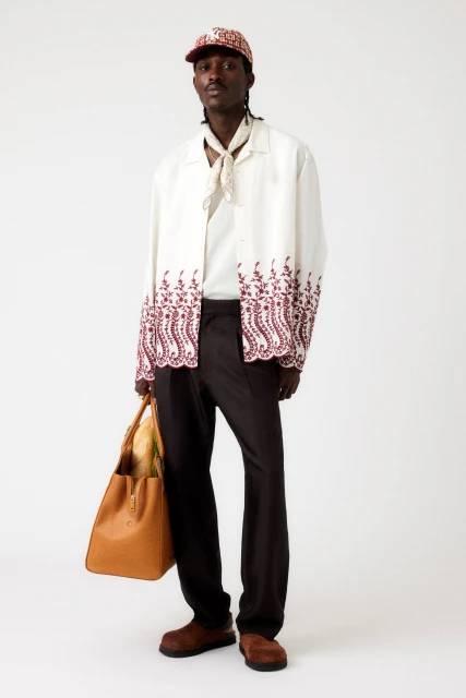 Full-length studio shot of a Black male model wearing a white button-down shirt with brown trim, dark pants, cap, and carrying a tan leather tote bag.