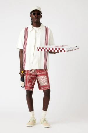 Stylish dark-skinned male model holds pizza and wine, showcasing patterned shorts, shirt, hat, and sunglasses against white.