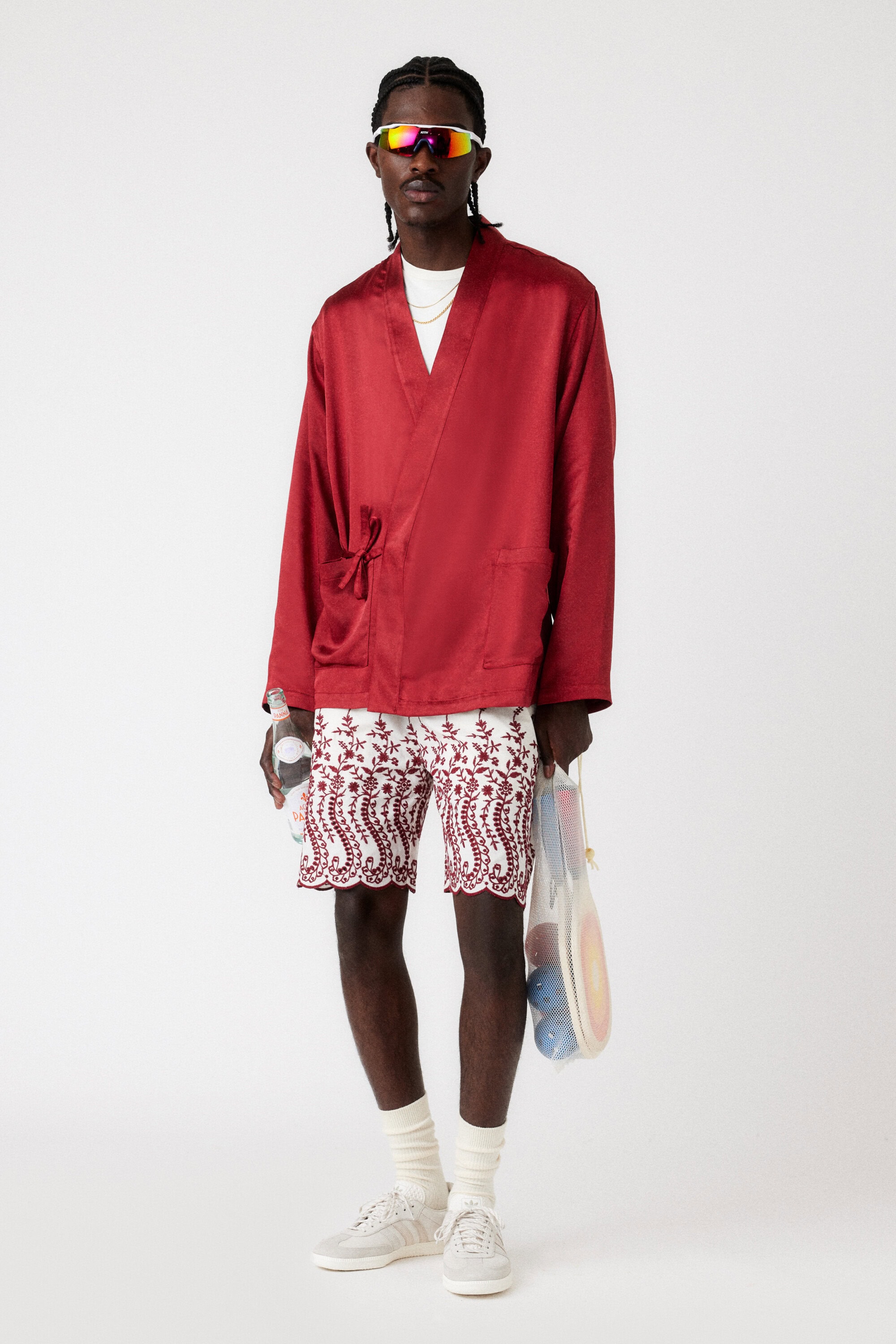 Adult male fashion model poses in a red satin robe, patterned shorts, socks, and sneakers against a white backdrop in a studio setting.