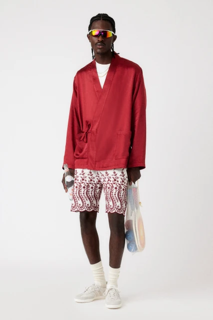 Adult male fashion model poses in a red satin robe, patterned shorts, socks, and sneakers against a white backdrop in a studio setting.