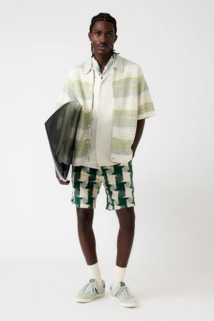 A Black man stands in a photo studio, wearing a button-down shirt, patterned shorts, socks, and sneakers, while holding a portfolio.