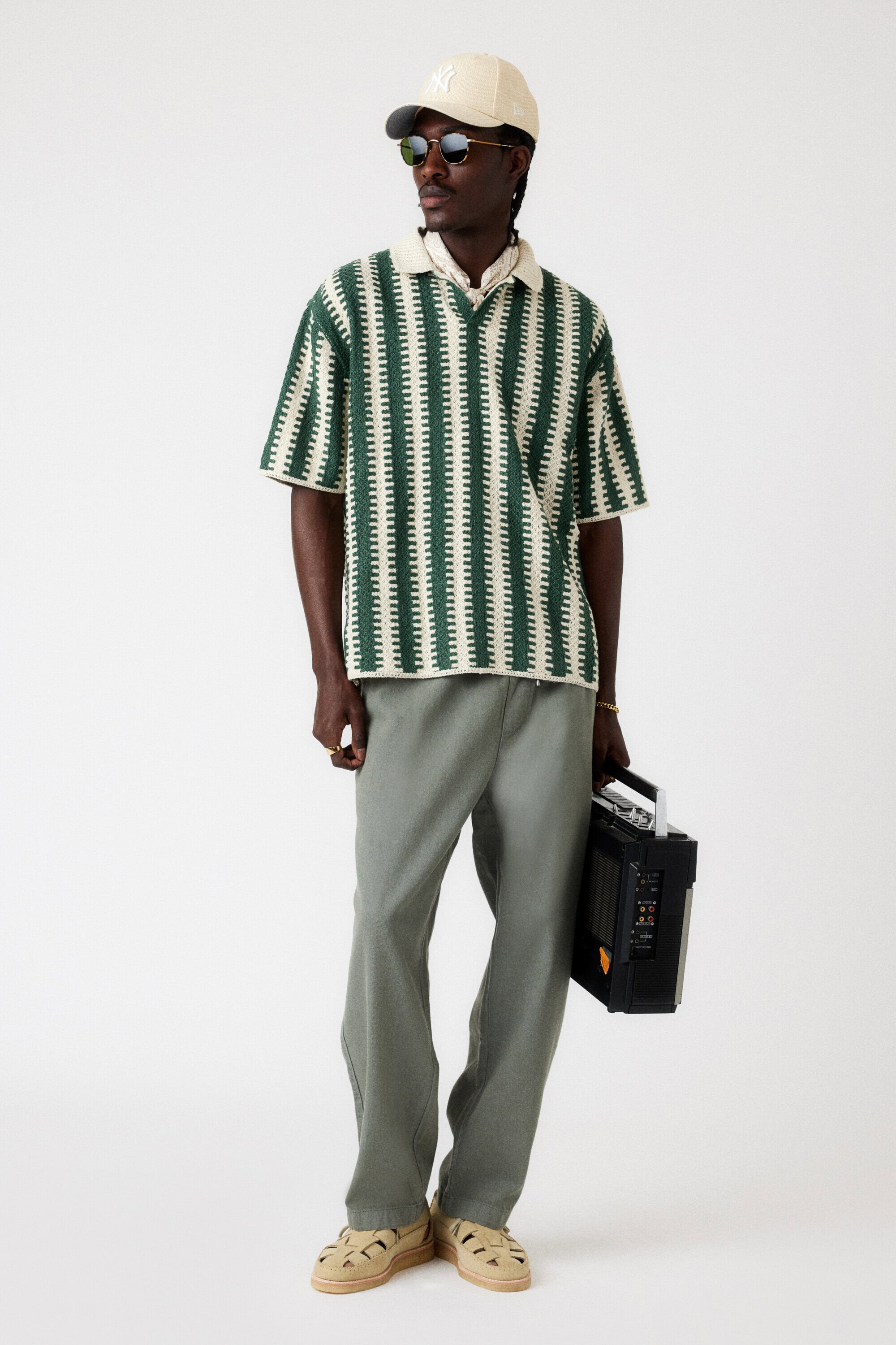 A dark-skinned man in a patterned polo, cap, and pants stands in a white studio, holding a black boombox in a fashion shot.