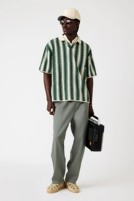 A dark-skinned man in a patterned polo, cap, and pants stands in a white studio, holding a black boombox in a fashion shot.