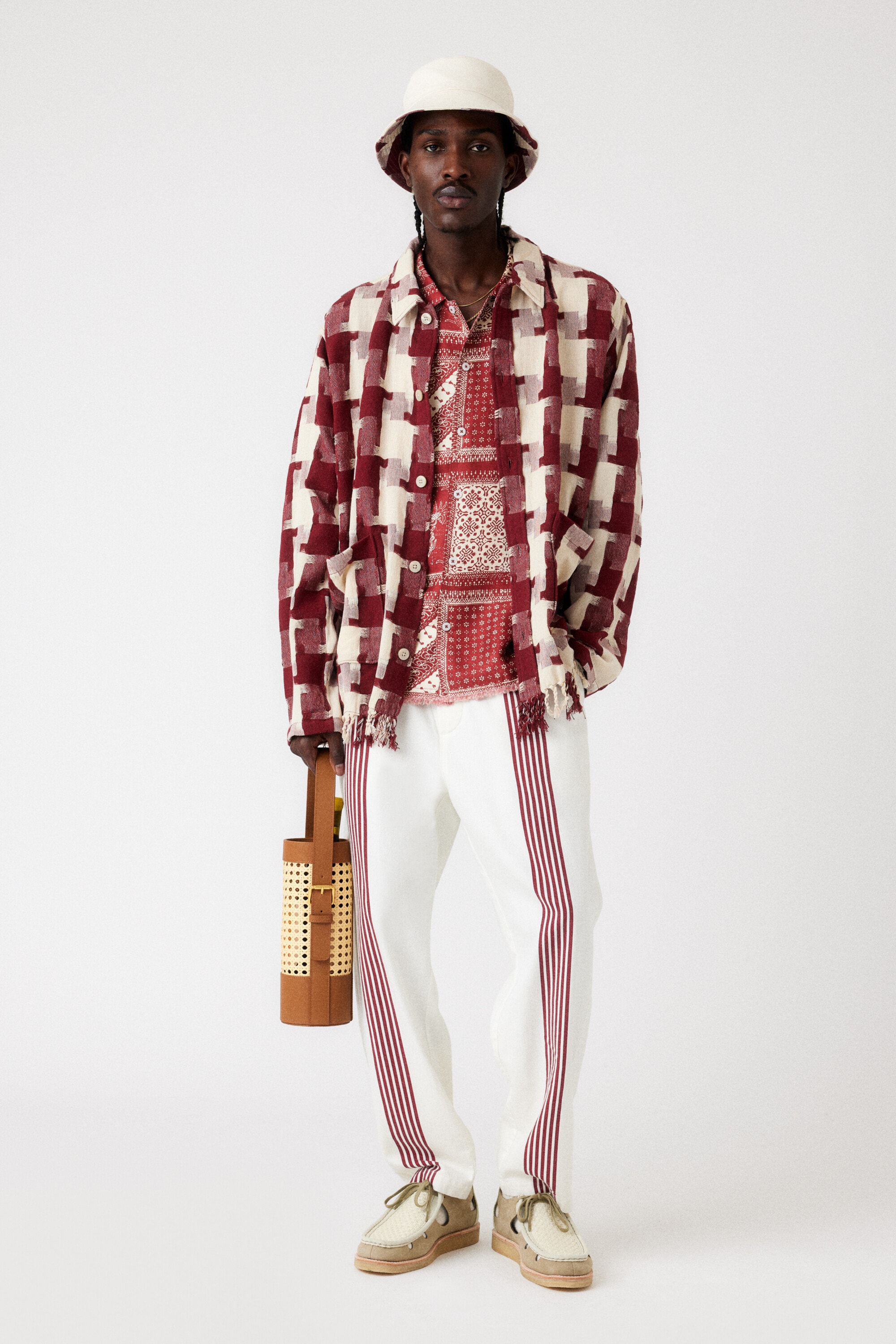 Adult male fashion model poses in a patterned shirt and trousers with a bucket hat, holding a wicker-handled water-bottle carrier.