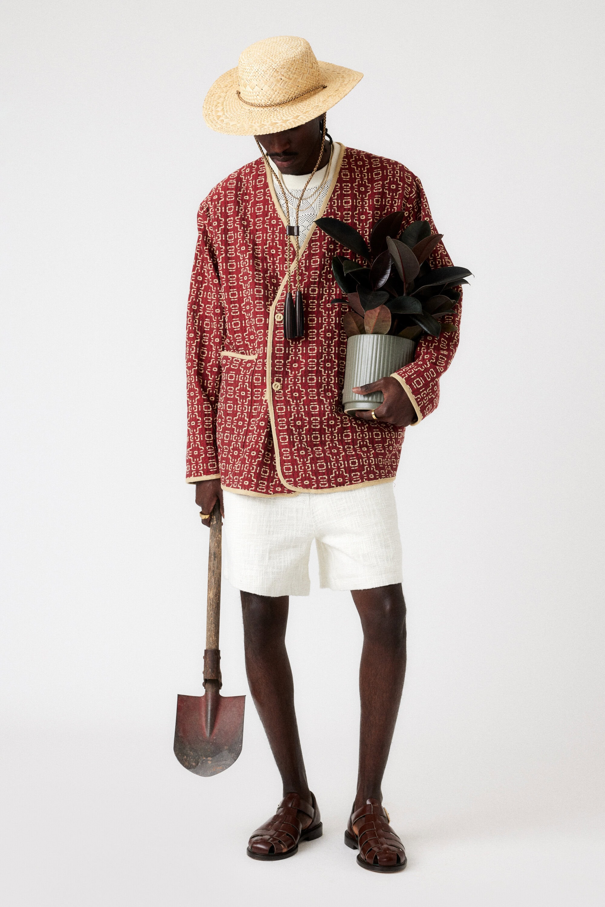 A dark-skinned male model stands in a studio wearing a patterned jacket, shorts, and a straw hat, holding a potted plant and shovel.
