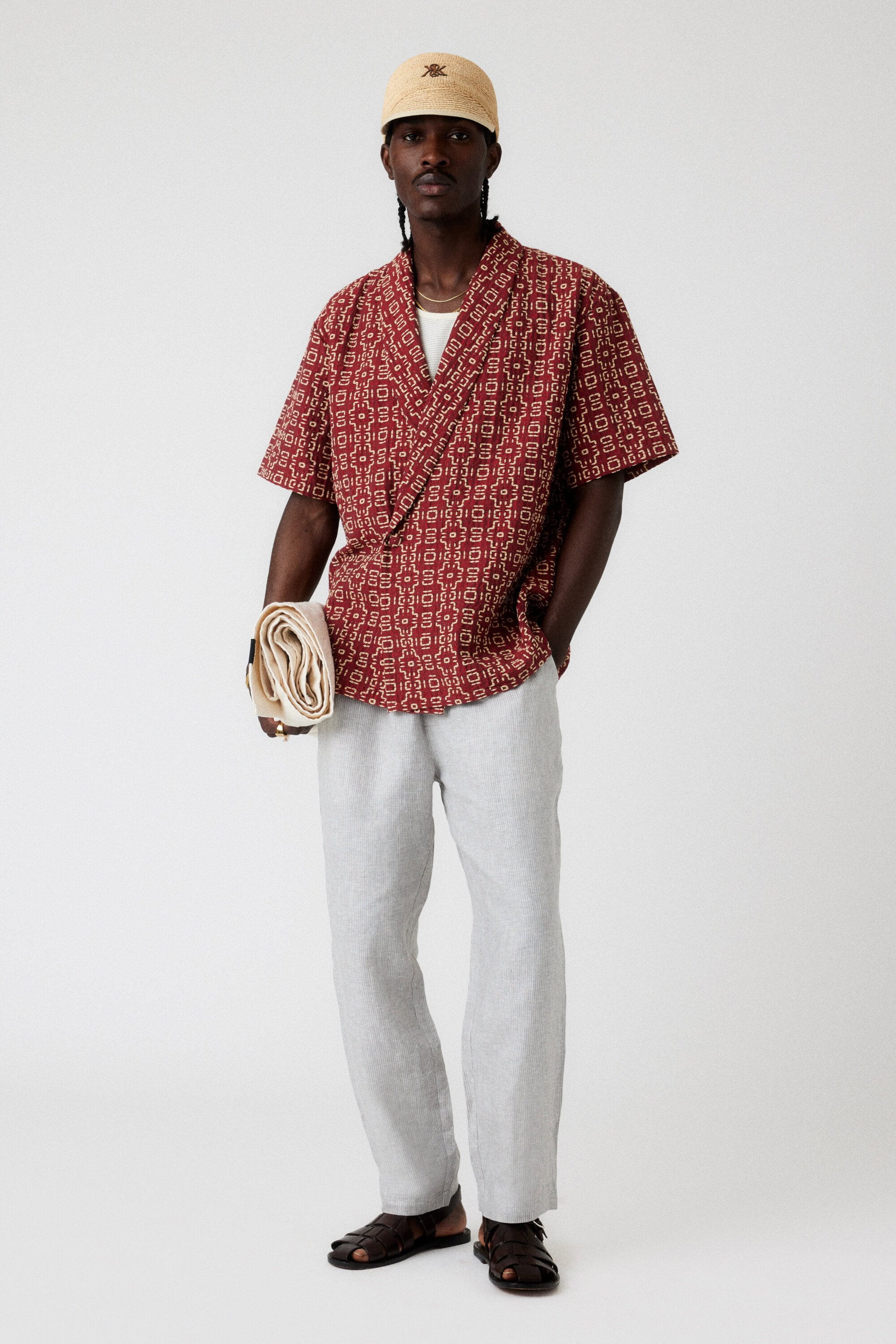 A full shot of a Black man in a patterned shirt, linen pants, and a cap, casually poses in a photo studio with neutral lighting.