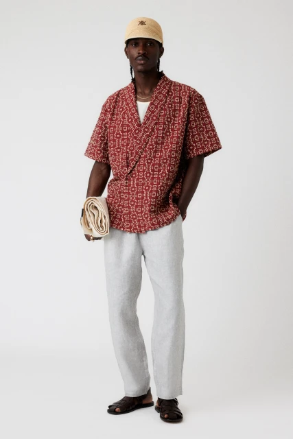A full shot of a Black man in a patterned shirt, linen pants, and a cap, casually poses in a photo studio with neutral lighting.