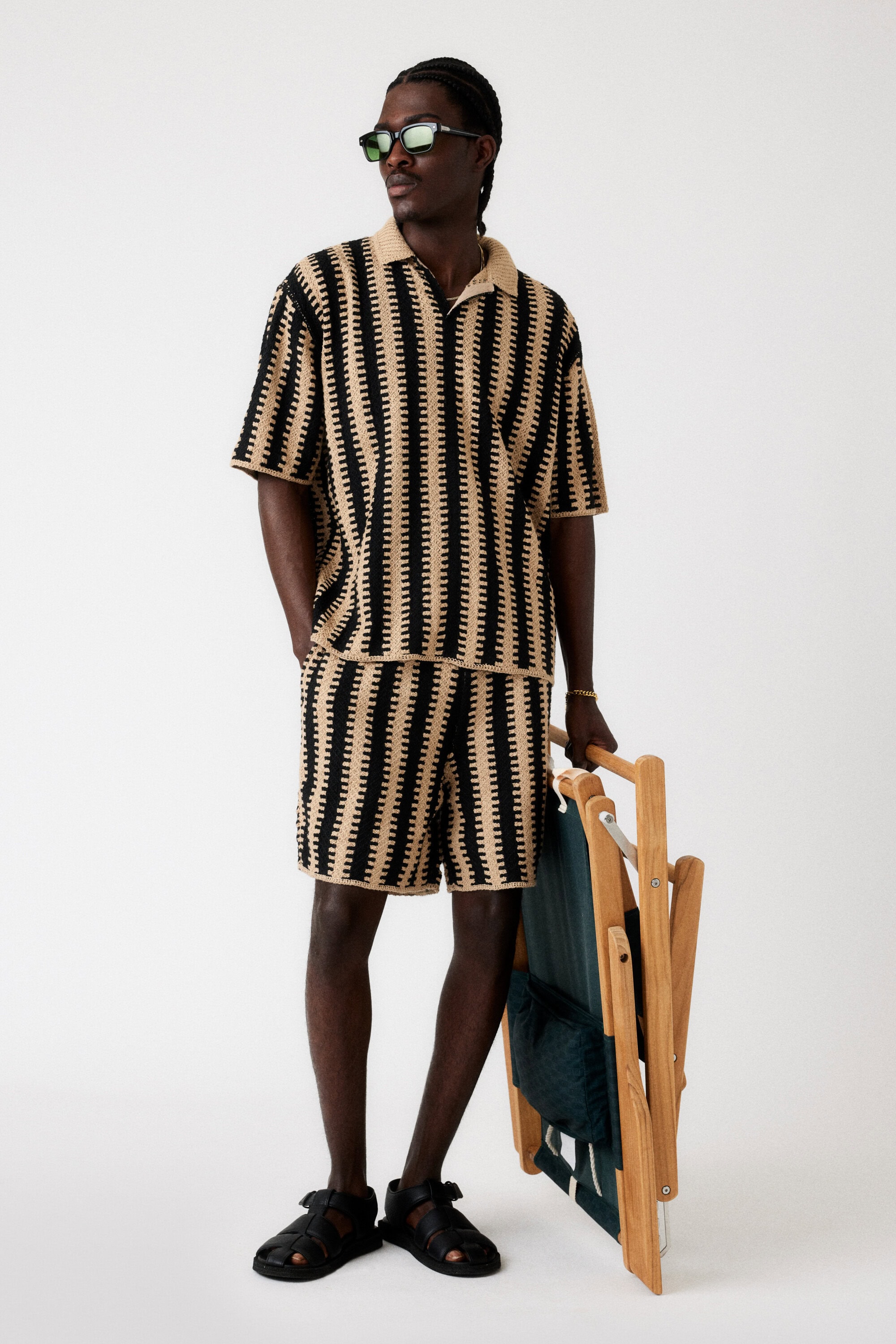 An adult Black male in a matching beige and black knit polo shirt and shorts holds a folded teal-and-wood beach chair in a studio shot.