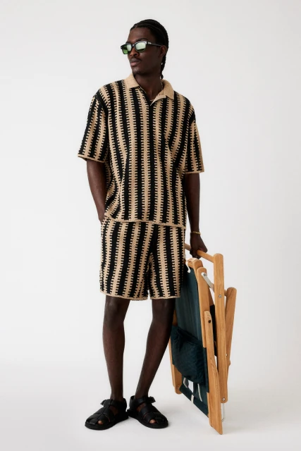 An adult Black male in a matching beige and black knit polo shirt and shorts holds a folded teal-and-wood beach chair in a studio shot.