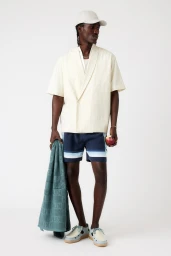 A man wearing a cream quilted short-sleeve shirt, blue striped shorts, and a cap, holding a rolled teal towel and an apple.