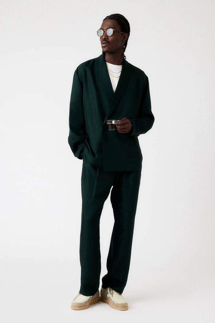 Man wearing a dark green suit jacket and trousers holding a small glass against a plain white background