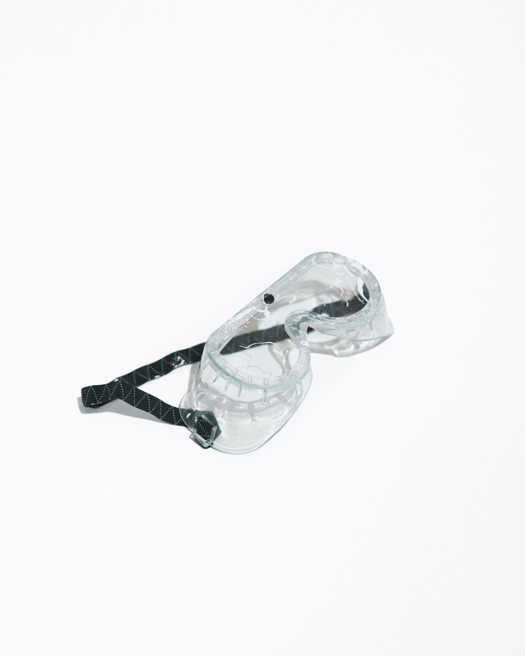 Clear protective eyewear with a dark green adjustable band lies on a clean white backdrop, casting a soft shadow. Safety and protection.