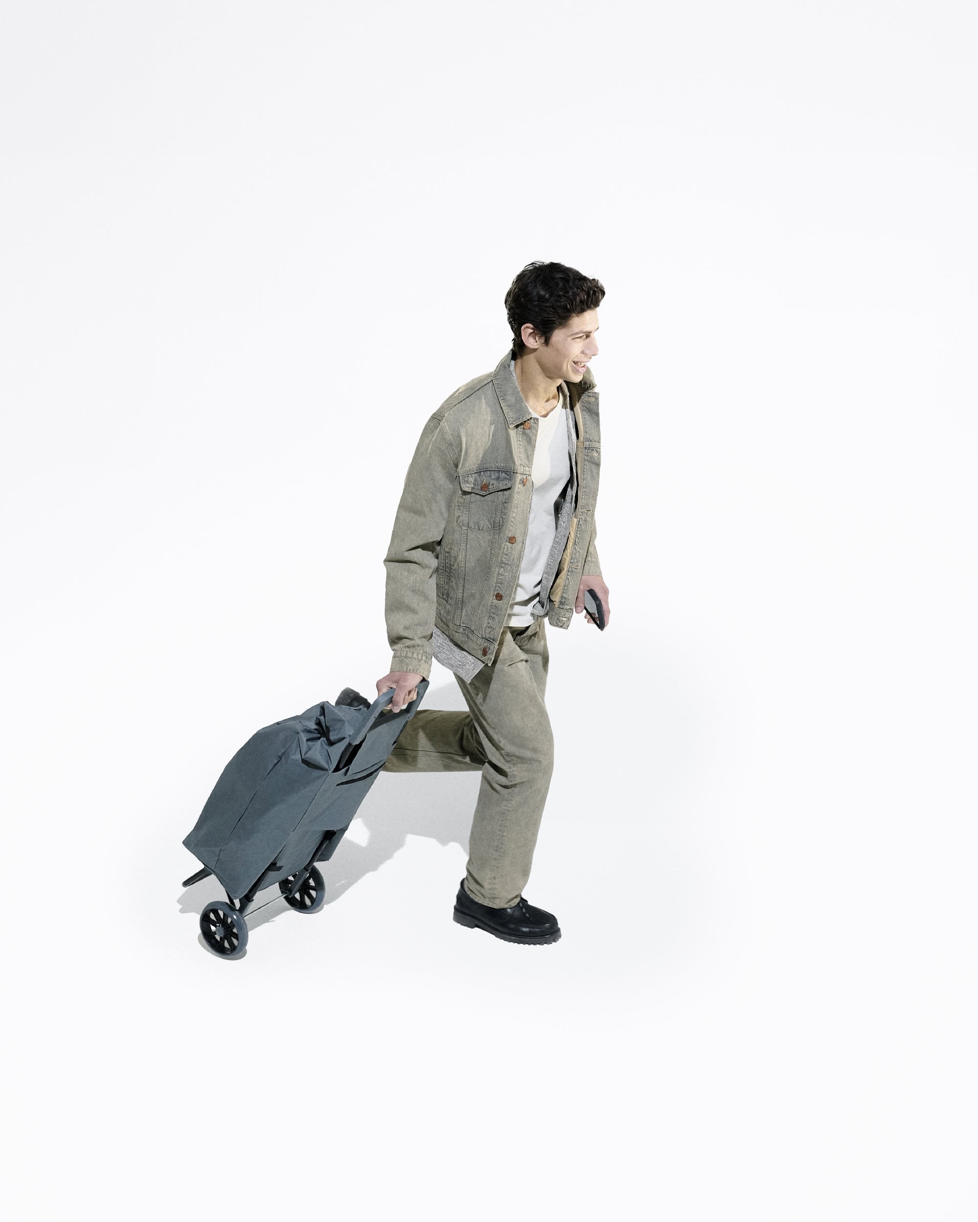 A young adult man walking with a rolling suitcase against a plain white backdrop, wearing casual clothing and smiling, in a full shot.