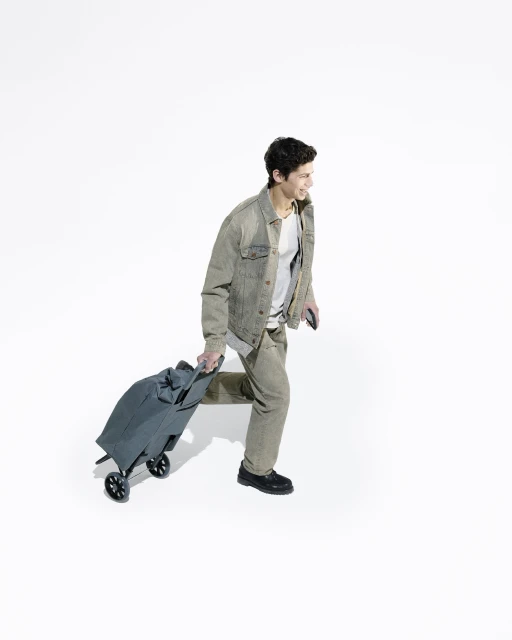 A young adult man walking with a rolling suitcase against a plain white backdrop, wearing casual clothing and smiling, in a full shot.