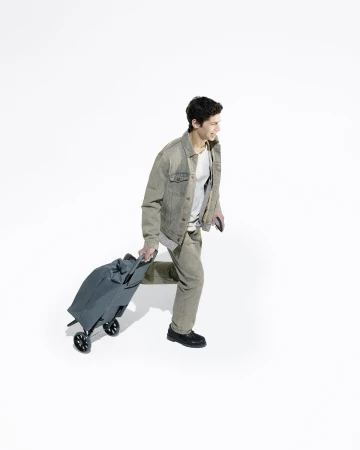 A young adult man walking with a rolling suitcase against a plain white backdrop, wearing casual clothing and smiling, in a full shot.