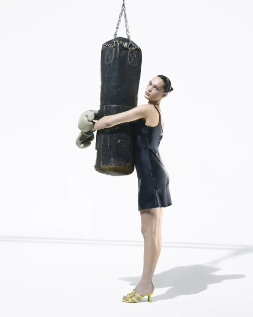 Woman in a dress and boxing gloves embraces a punching bag in a studio with a white background. The scene is lit with bright light.