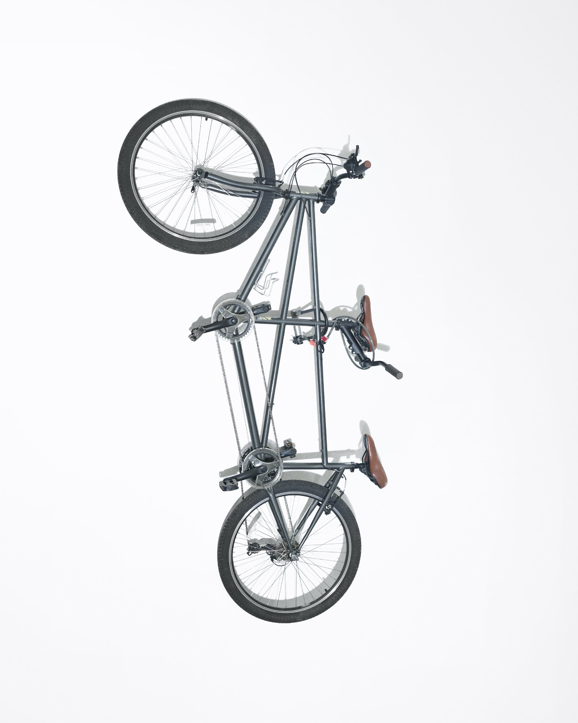 Studio shot of a gray tandem bicycle, angled vertically, against a bright white background. Wheels, seats, and frame are clearly visible.