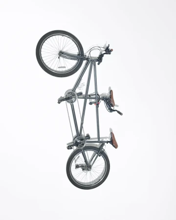 Studio shot of a gray tandem bicycle, angled vertically, against a bright white background. Wheels, seats, and frame are clearly visible.