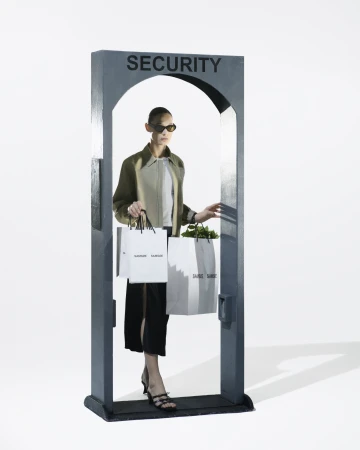 A fashion model is walking through a grey metal detector-like structure with shopping bags in hand against a plain white background.