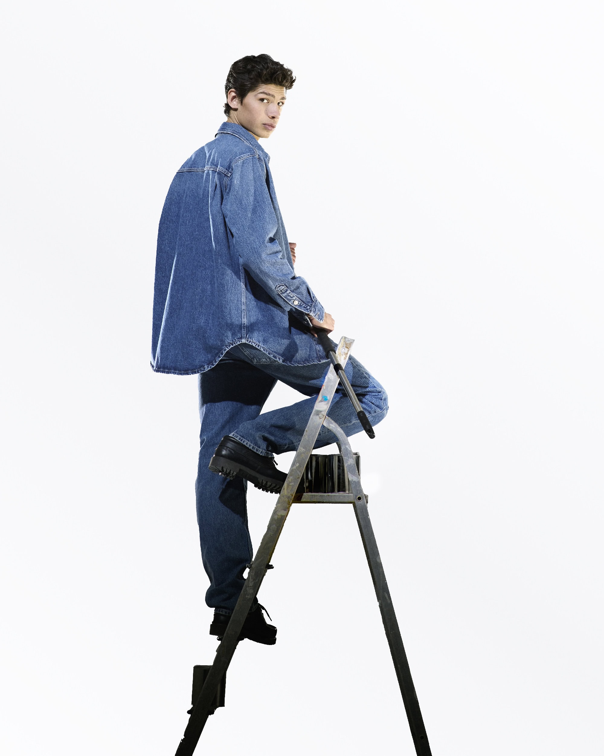 Young man in a denim jacket and jeans stands on a silver step ladder against a solid white backdrop in a full shot, looking back.