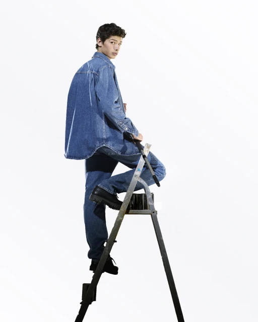 Young man in a denim jacket and jeans stands on a silver step ladder against a solid white backdrop in a full shot, looking back.