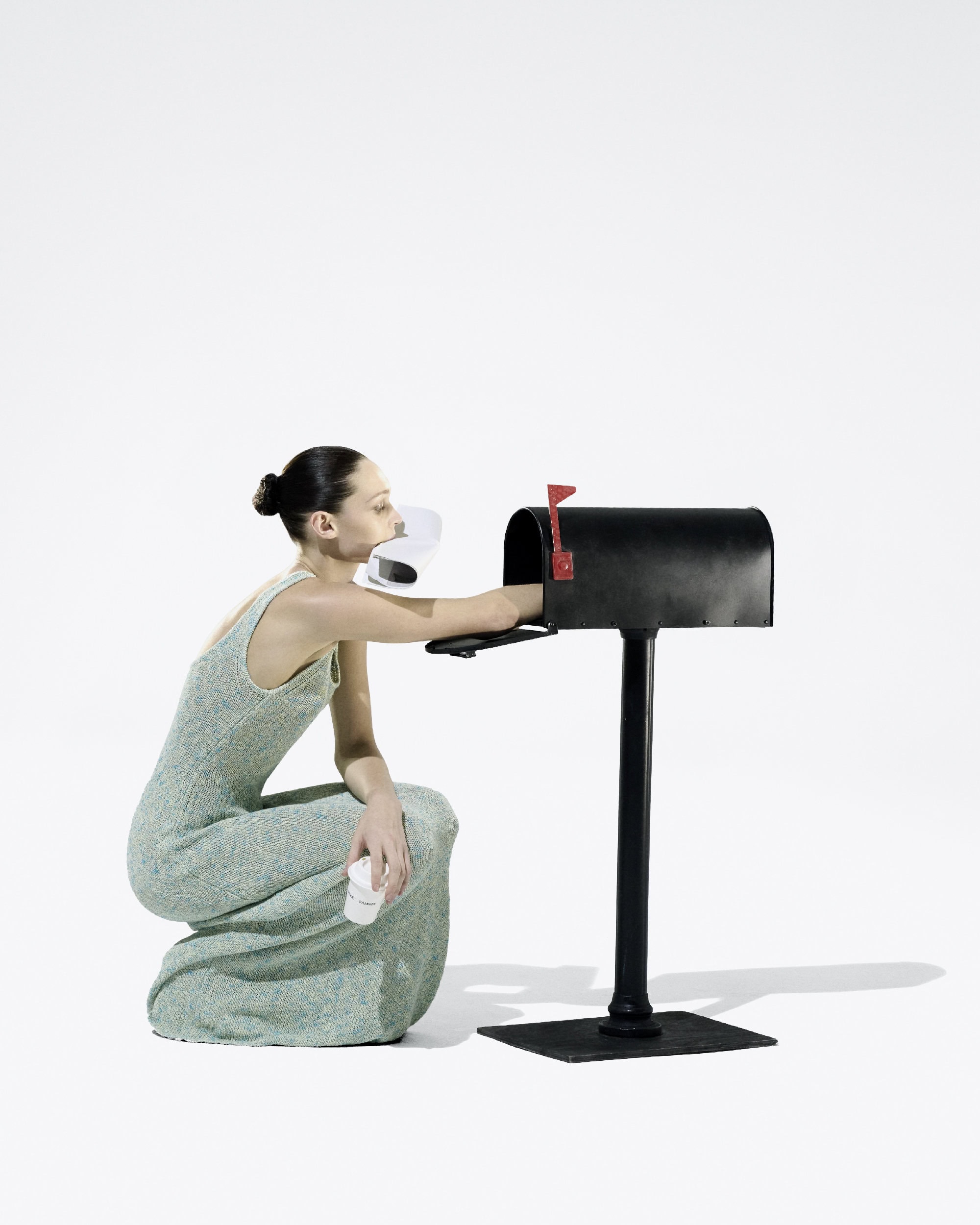 Woman in a long dress squats next to a black mailbox, with a letter in her mouth and another in her hand, against a white background.