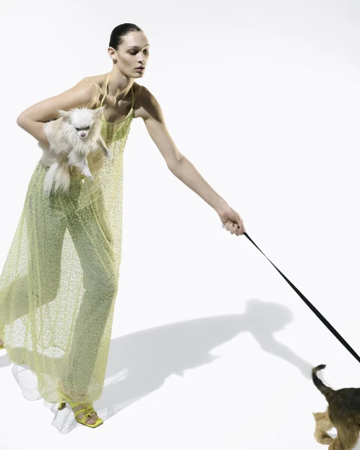 Woman in yellow dress holds a white fluffy dog and walks another dog on a leash in a fashion studio setting against a white backdrop.