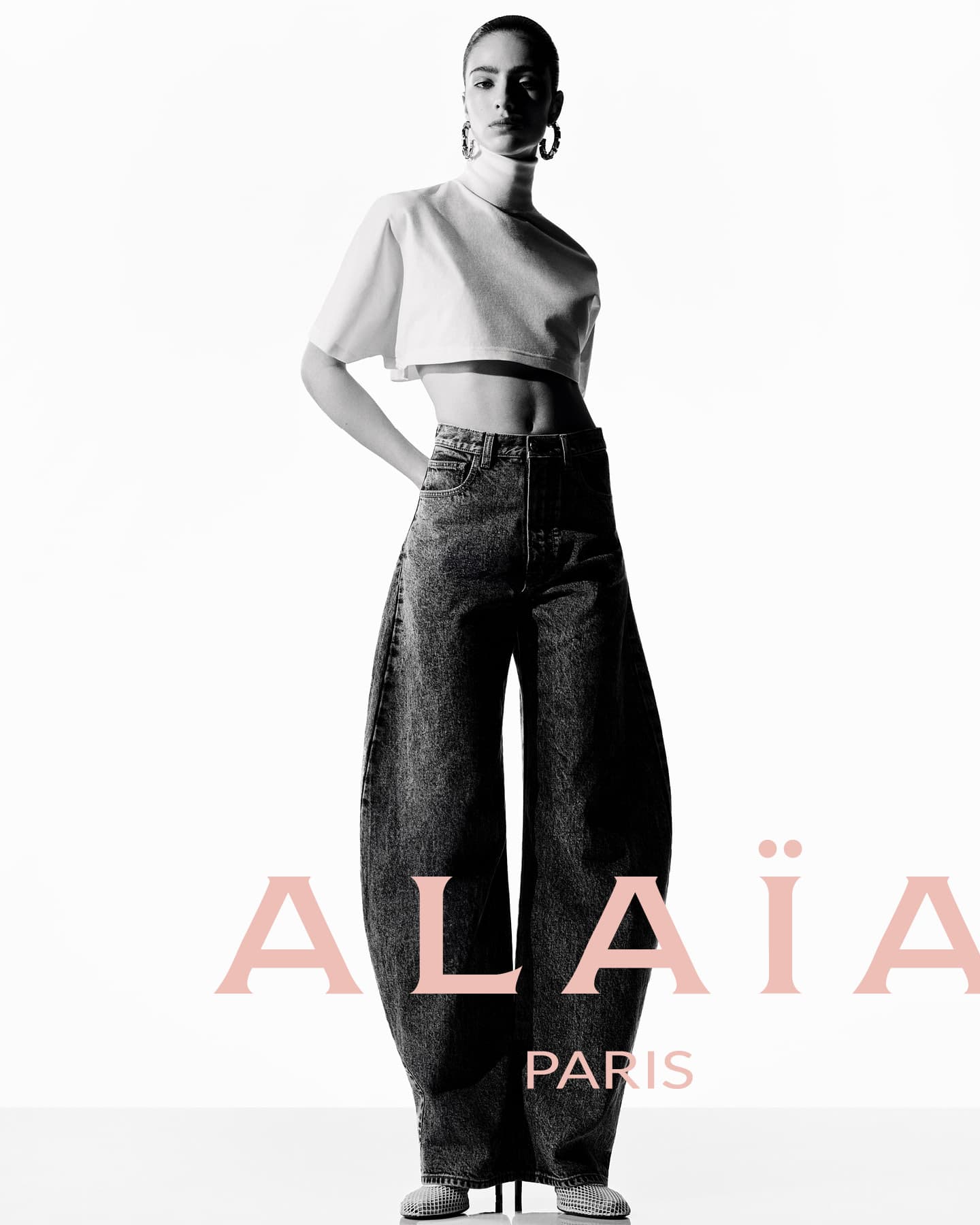 A woman poses in a fashion shot wearing a white crop top and oversized denim pants against a bright studio backdrop with brand logo.