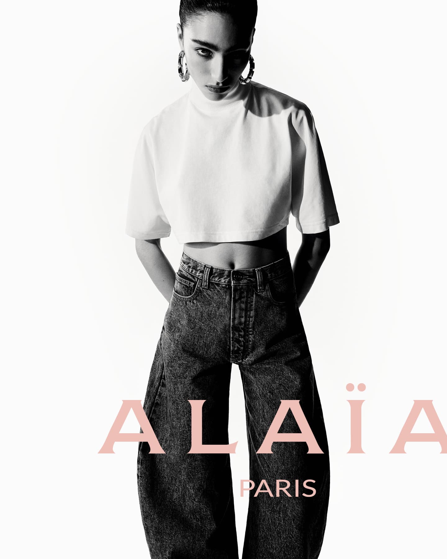 Black and white fashion shot of a woman in a white crop top and dark jeans standing against a bright background, logo visible.
