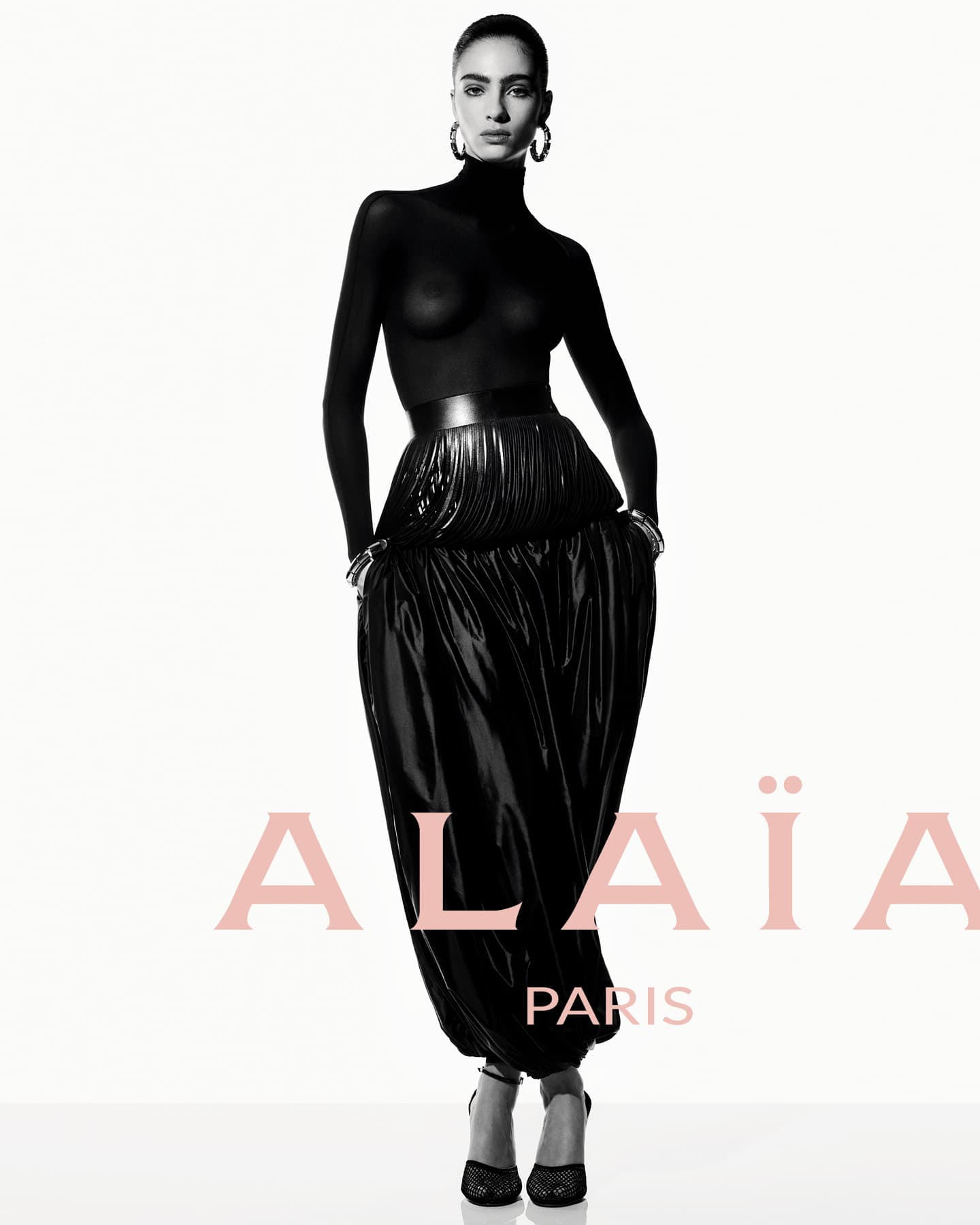 Fashion editorial shot in studio featuring a slender adult woman in a black turtleneck and dramatic black skirt and high heels.