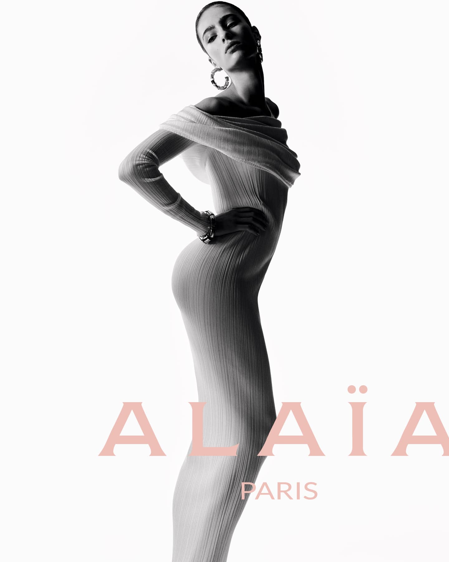 Fashion shot of a female model in a full-length off-shoulder dress with long sleeves, posing in a photo studio against a white backdrop.