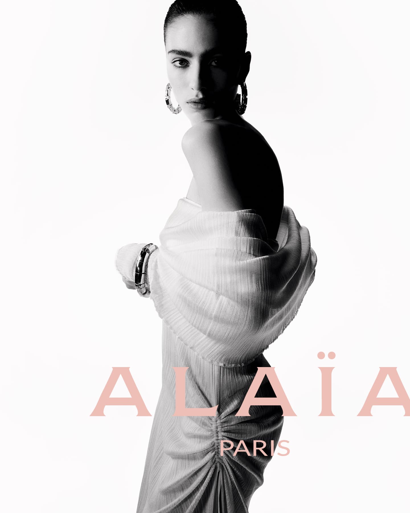 Elegant portrait of a fashion model in a white dress and hoop earrings posing in front of a stark white background with Alaïa branding.