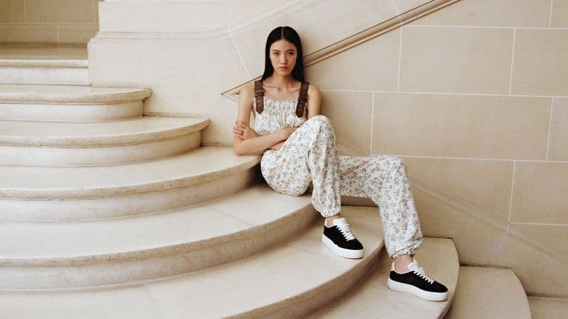 Asian woman in floral jumpsuit and black sneakers sits on cream-colored staircase, gazing at the camera, in a neutral indoor setting.