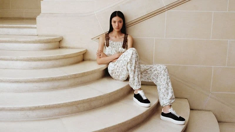 Asian woman in floral jumpsuit and black sneakers sits on cream-colored staircase, gazing at the camera, in a neutral indoor setting.