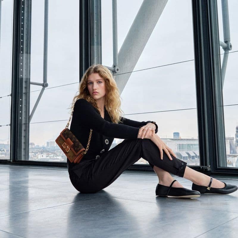 A blonde fashion model poses while sitting indoors in black attire and Mary Jane shoes next to a Louis Vuitton handbag.