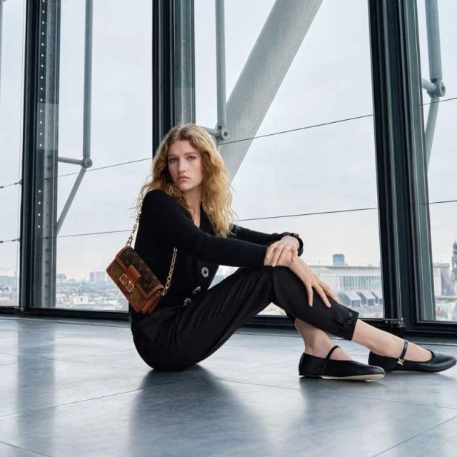 A blonde fashion model poses while sitting indoors in black attire and Mary Jane shoes next to a Louis Vuitton handbag.
