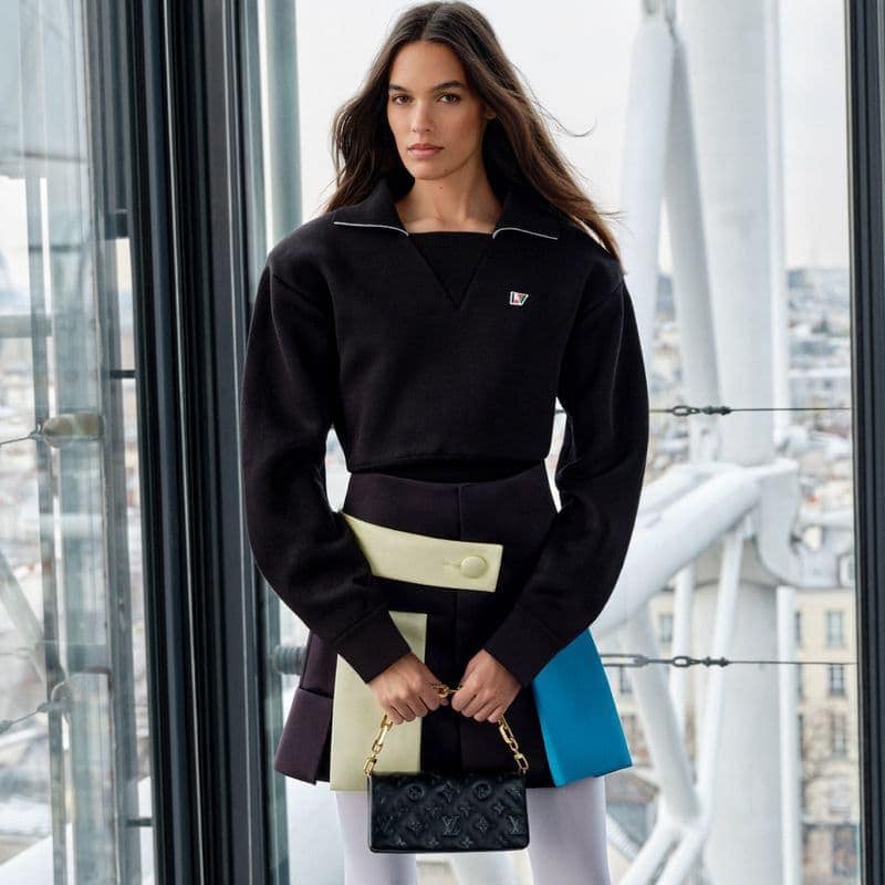 A fashionable adult woman poses in front of a modern glass-walled building, wearing a black top and colorful skirt, and holding a small black purse.