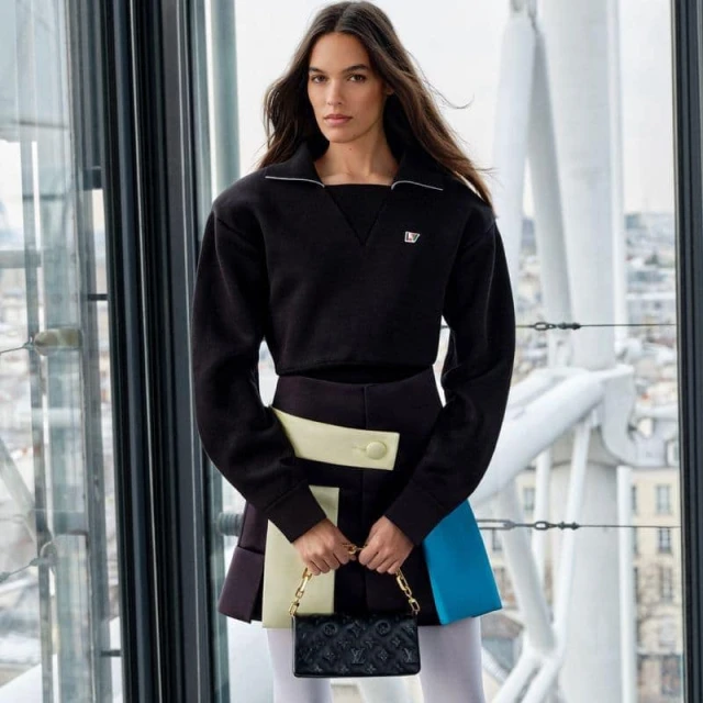 A fashionable adult woman poses in front of a modern glass-walled building, wearing a black top and colorful skirt, and holding a small black purse.