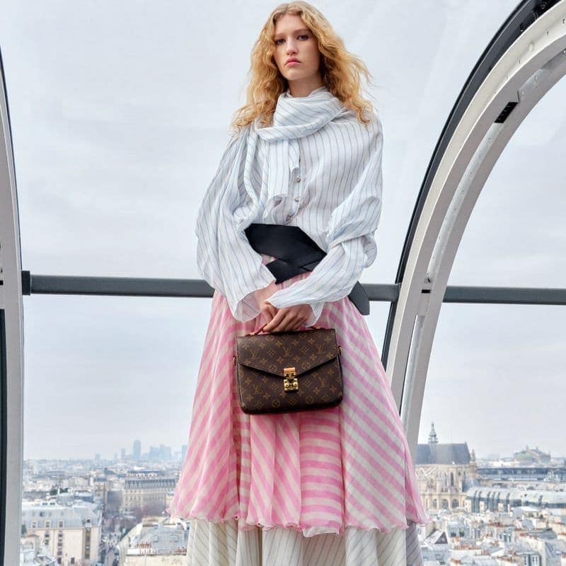 Fashionable woman in a white striped shirt and pink striped skirt holding a brown Louis Vuitton handbag in front of a city skyline.