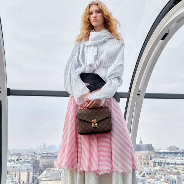Fashionable woman in a white striped shirt and pink striped skirt holding a brown Louis Vuitton handbag in front of a city skyline.