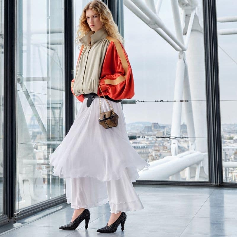 Fashion shot of a woman in a beige and orange jacket, layered white skirt, and snakeskin heels, standing inside with city view backdrop.