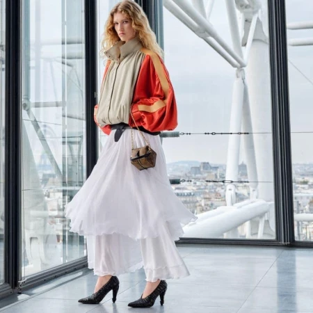 Fashion shot of a woman in a beige and orange jacket, layered white skirt, and snakeskin heels, standing inside with city view backdrop.