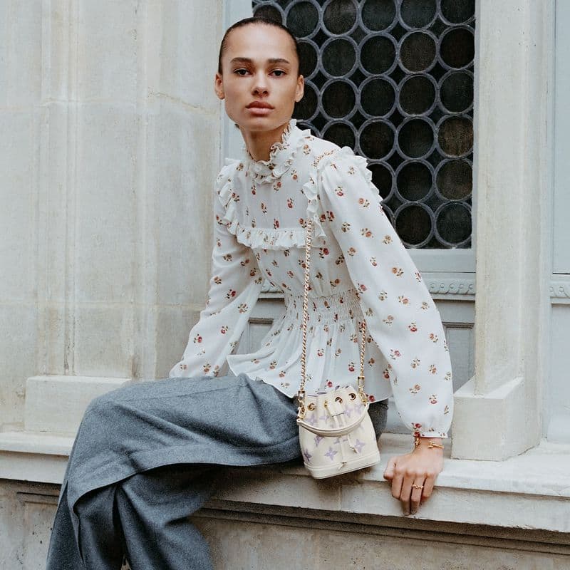 A woman in a floral blouse and grey pants sits on a ledge, a small bucket bag with a gold chain strap hangs from her shoulder.