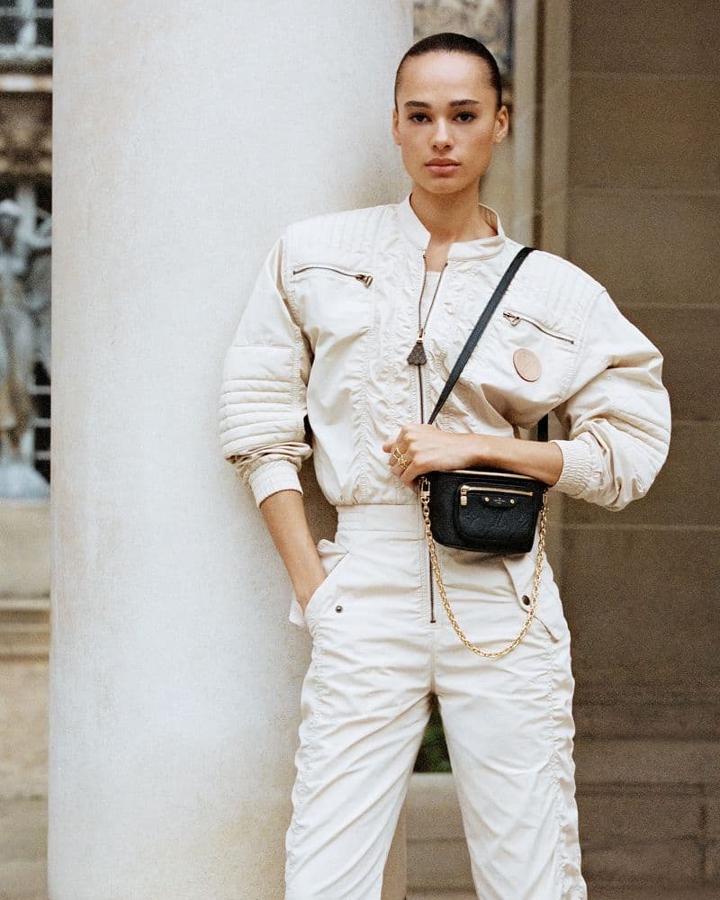 A woman in a cream-colored jumpsuit poses with a black crossbody bag in front of a stone building with classical architecture.