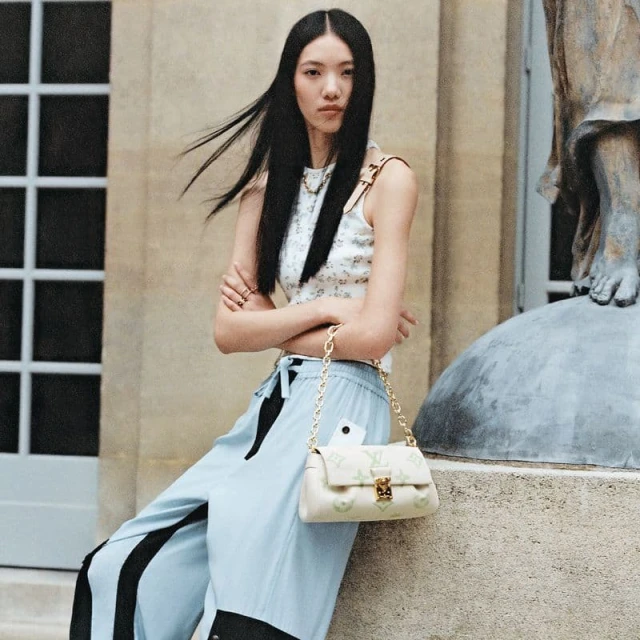Elegant Asian woman posing outdoors in a floral top, wide-leg pants, and a stylish handbag with a gold chain strap, near a stone statue.