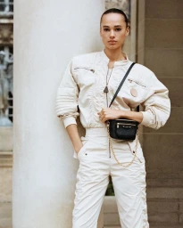 Model wearing an off-white zip-up jacket and matching trousers, posing with a small black crossbody bag against a large white column.