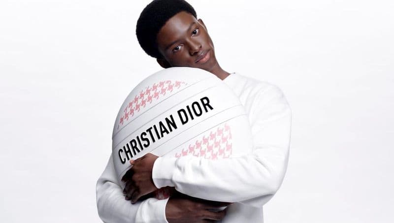 Portrait of a Black male in a white sweater, embracing a white circular object with a pink pattern and the text CHRISTIAN DIOR.