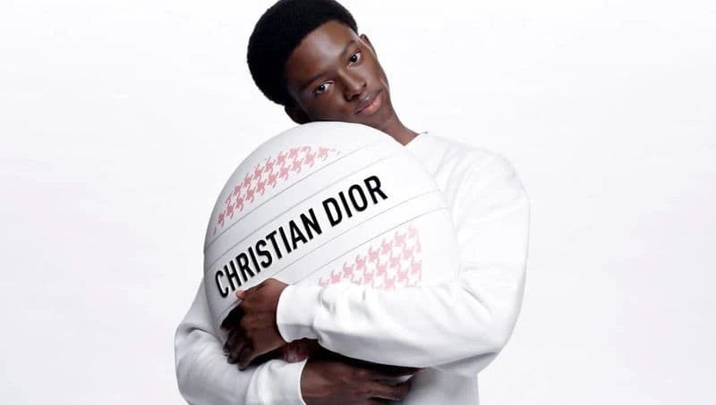 Portrait of a Black male in a white sweater, embracing a white circular object with a pink pattern and the text CHRISTIAN DIOR.